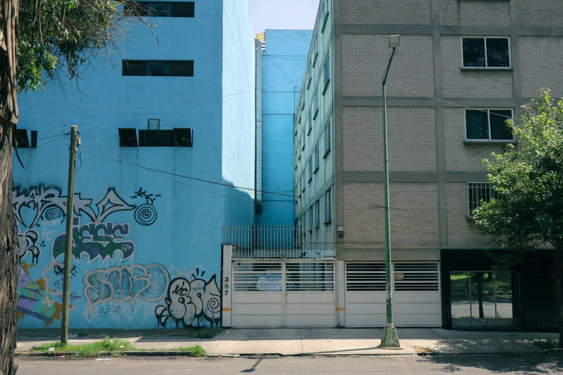 Urban street scene with graffiti on a blue wall, a brick building with several windows, utility poles, streetlights, and trees.