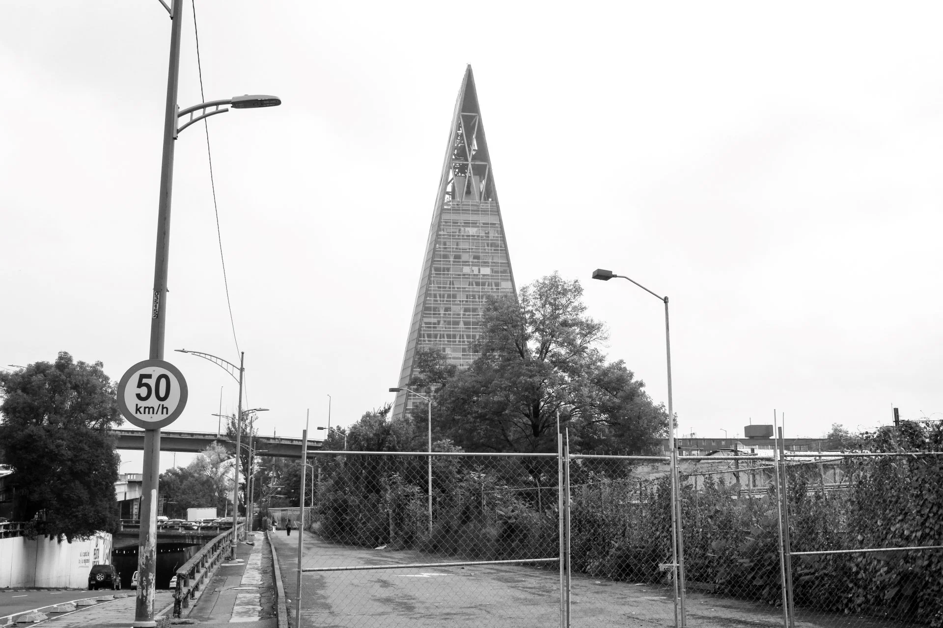 View of a street with a tall pyramid-shaped building in the background, streetlights, trees, a sidewalk, and a speed limit sign.