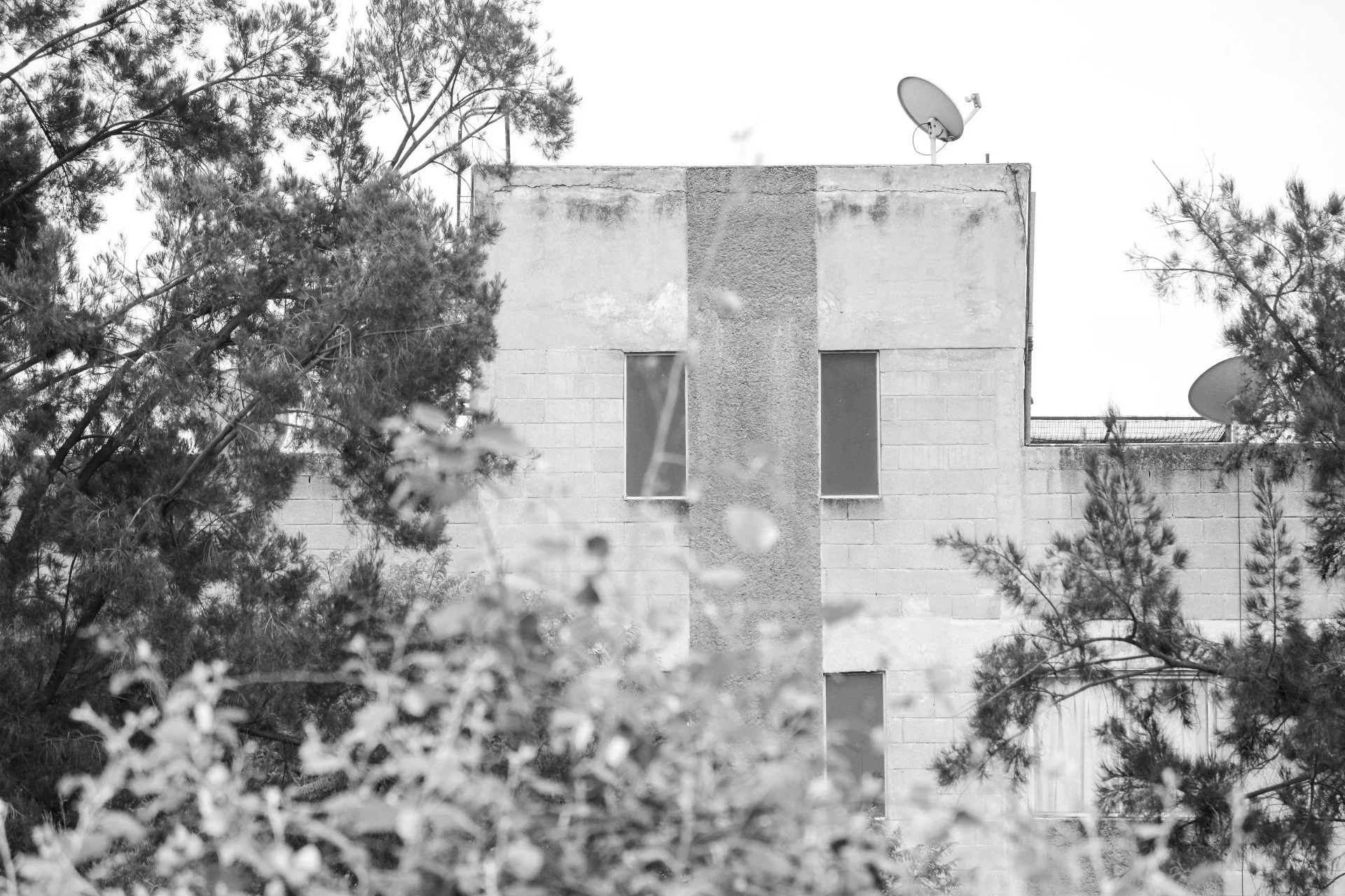 Black and white photo of the upper side of a building with trees around it. The building has two windows and two satellite dishes on the roof.