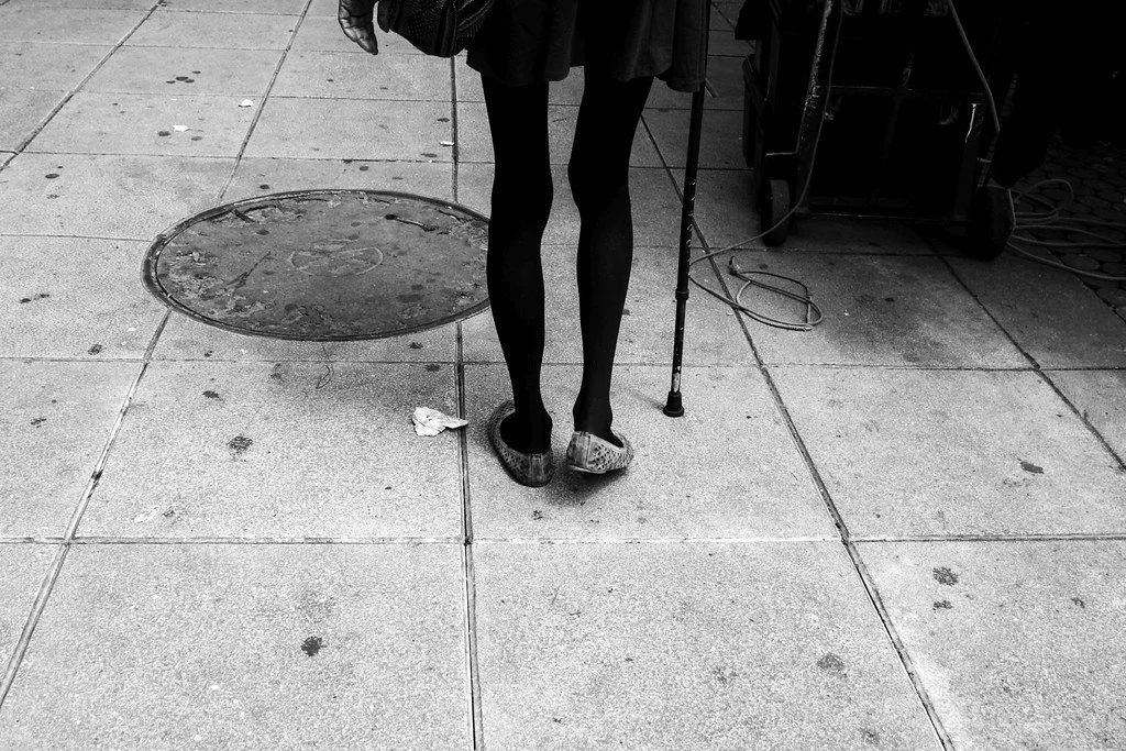 A person standing on a concrete sidewalk with a crutch and wearing slippers, black stockings, and a coat. There is a manhole cover and some scattered trash on the sidewalk.