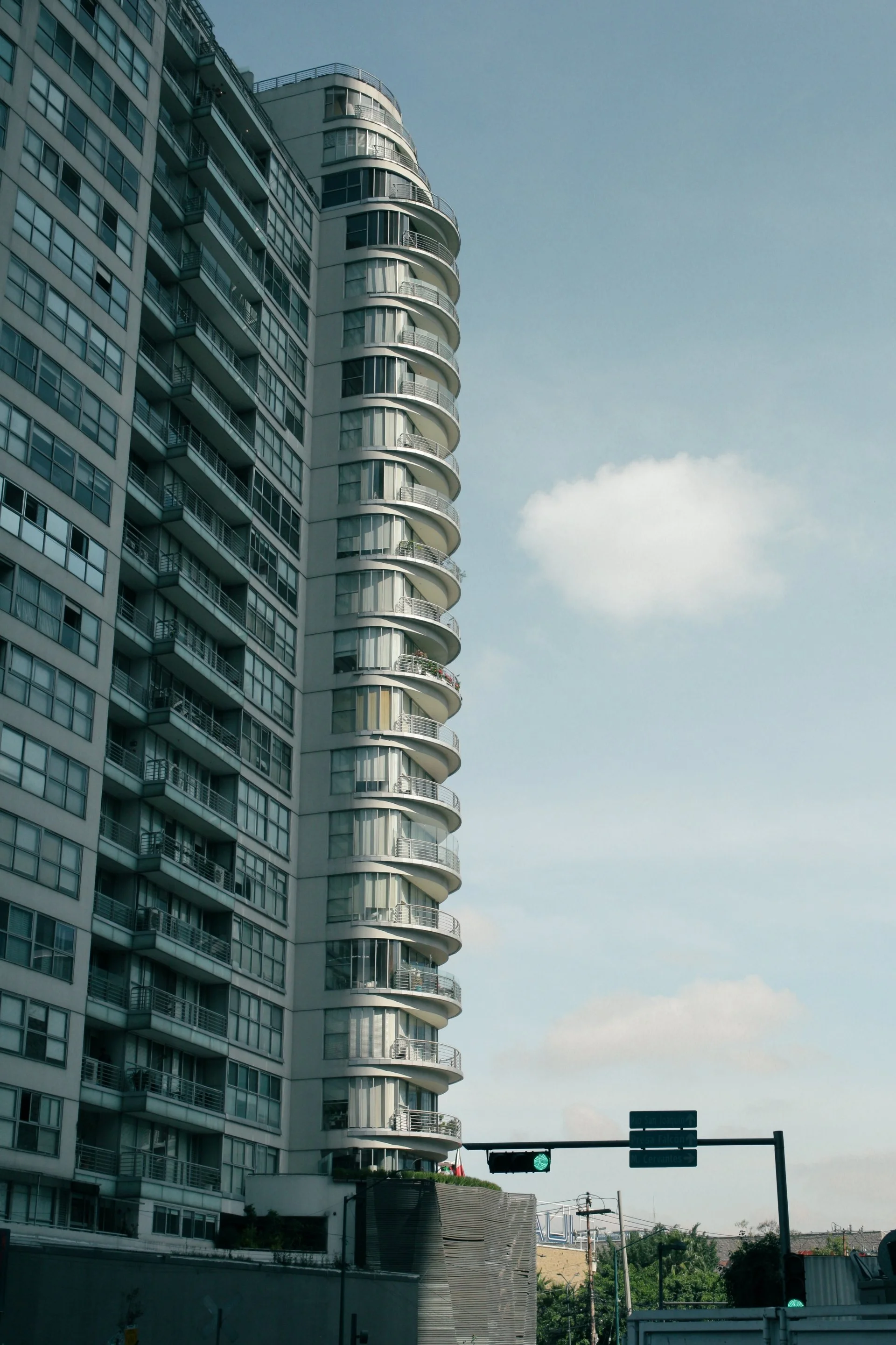 High-rise modern apartment building with curved balconies on a city street, traffic lights, and a blue sky with some clouds in the background.