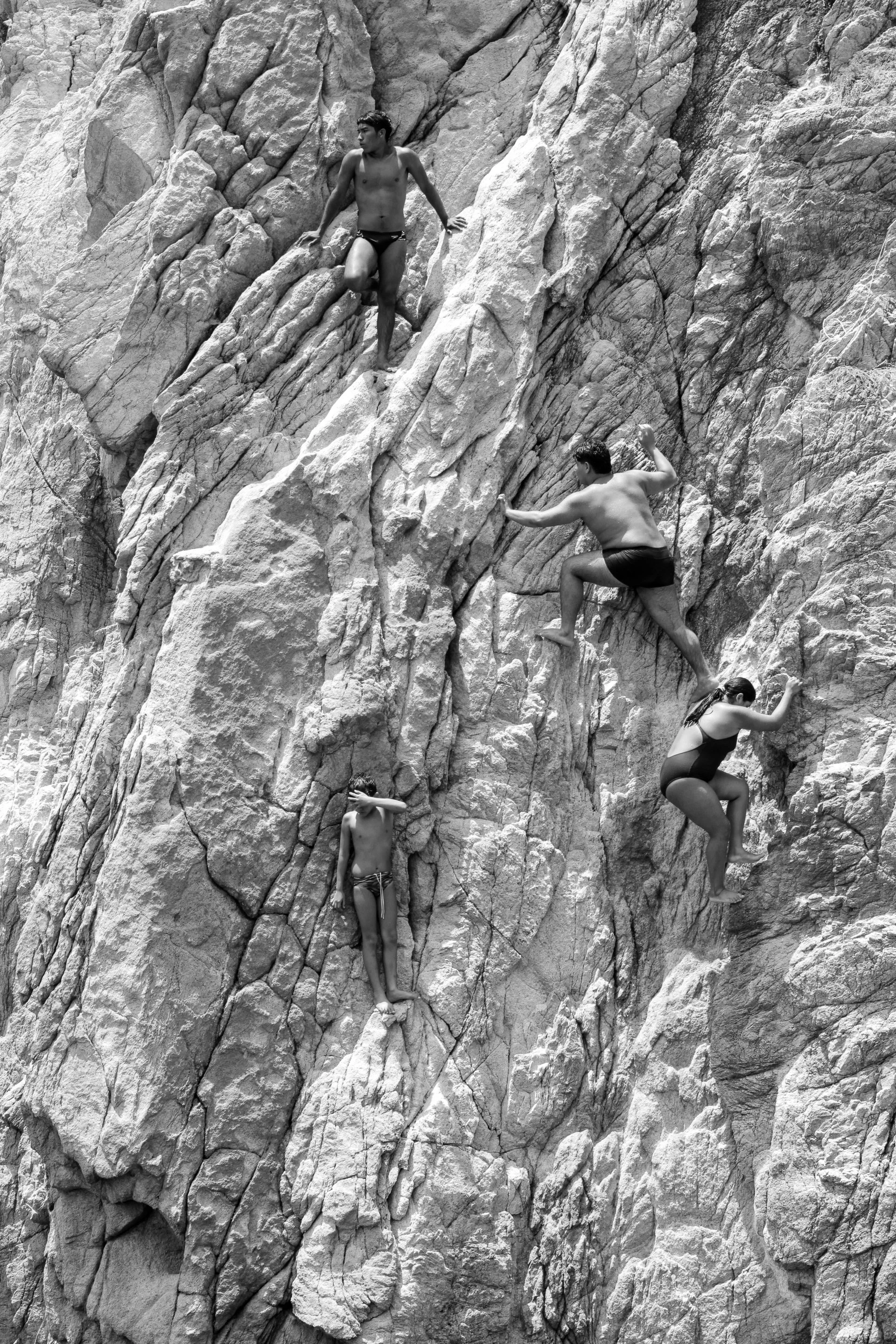 Black and white photo of four people rock climbing on a steep, rocky cliff. One male climber is higher up, wearing only swim trunks. Another male climber is on a lower level, also in swim trunks and reaching for a hold. Two female climbers are lower 