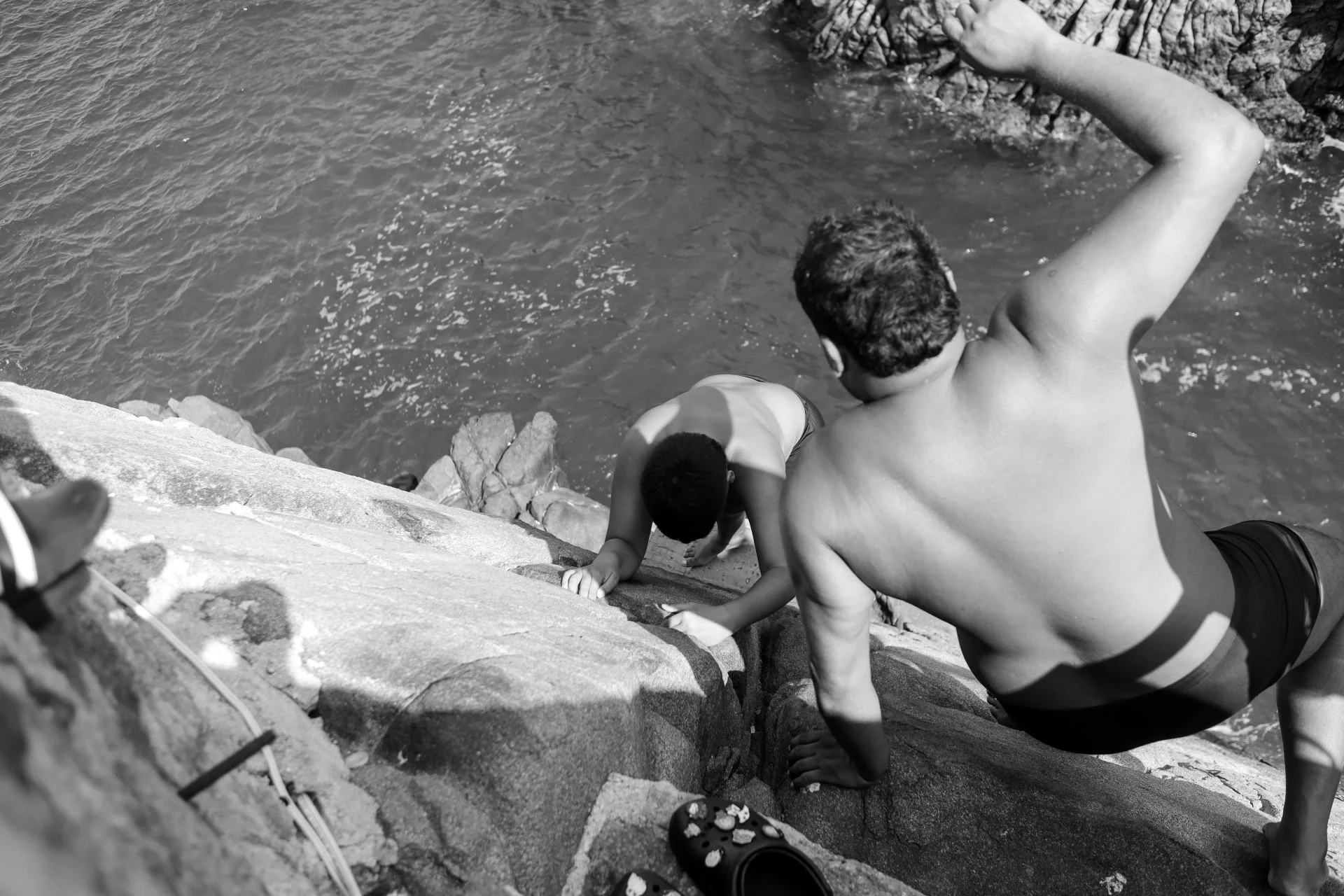 A man and a young boy, both shirtless, leaning over a rocky ledge by the water's edge. The man is reaching out with his right arm, and the boy is using both hands for support on the rocks. The water is calm, and part of the rocky shoreline is visible