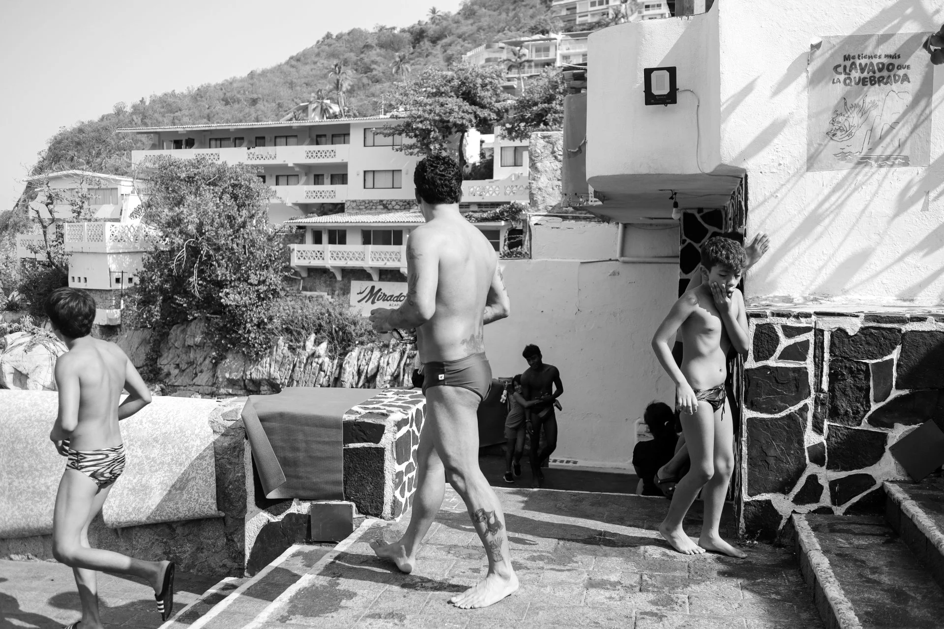 Group of children in swimsuits standing and playing near a stone structure outdoors, with buildings and a hillside in the background.