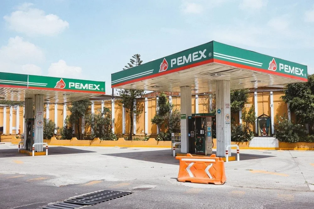 A Pemex gas station with two fueling islands, a black payment kiosk, and a large orange safety barrier in the foreground.
