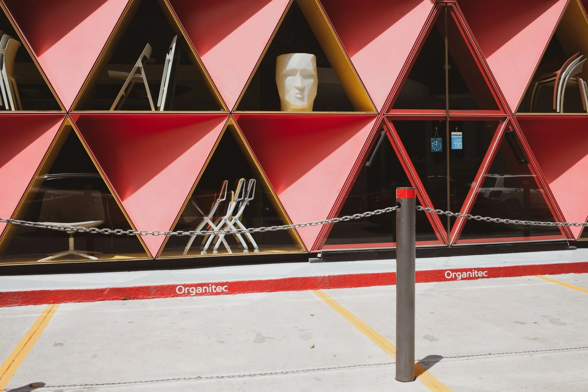 Storefront with red and black geometric triangular shelving units displaying chairs and a decorative mask, with a chain barrier and parking lot in front.