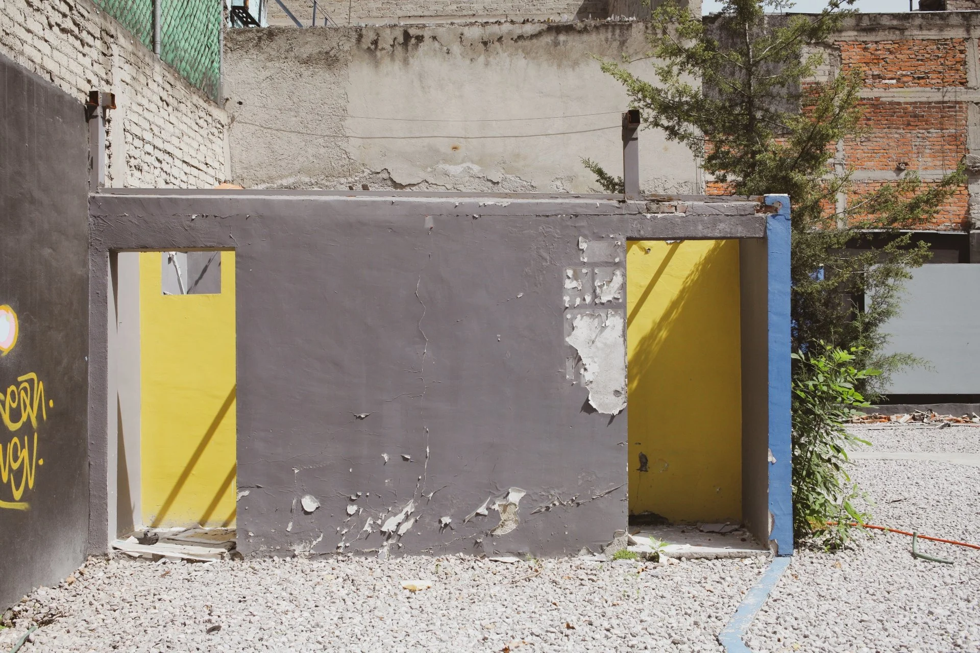 An exterior view of a small building with a yellow door and gray walls, in a state of disrepair. The building has peeling paint and cracks, with graffiti on one wall. Surrounding area is gravel with some greenery and trees, and other buildings in the
