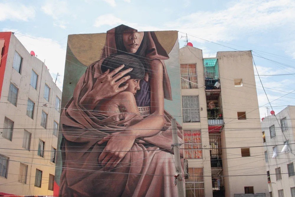Large mural on a building depicting a woman with a hood embracing a young girl, painted in realistic style, across city apartment buildings under a partly cloudy sky.