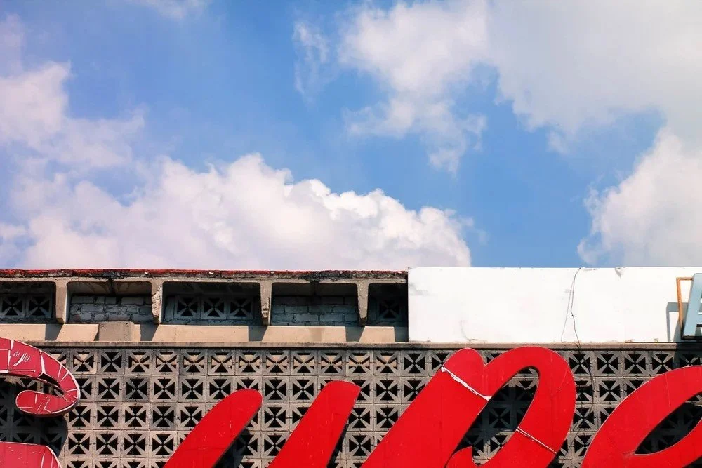 Part of a building with a decorative vented wall and large red sign lettering, against a blue sky with clouds.