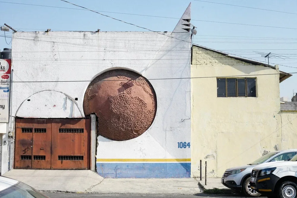 Building with mural of the moon in a craters phase, part of the building is painted white, and the other part is painted yellow with the number 1064. There are parked cars in front.