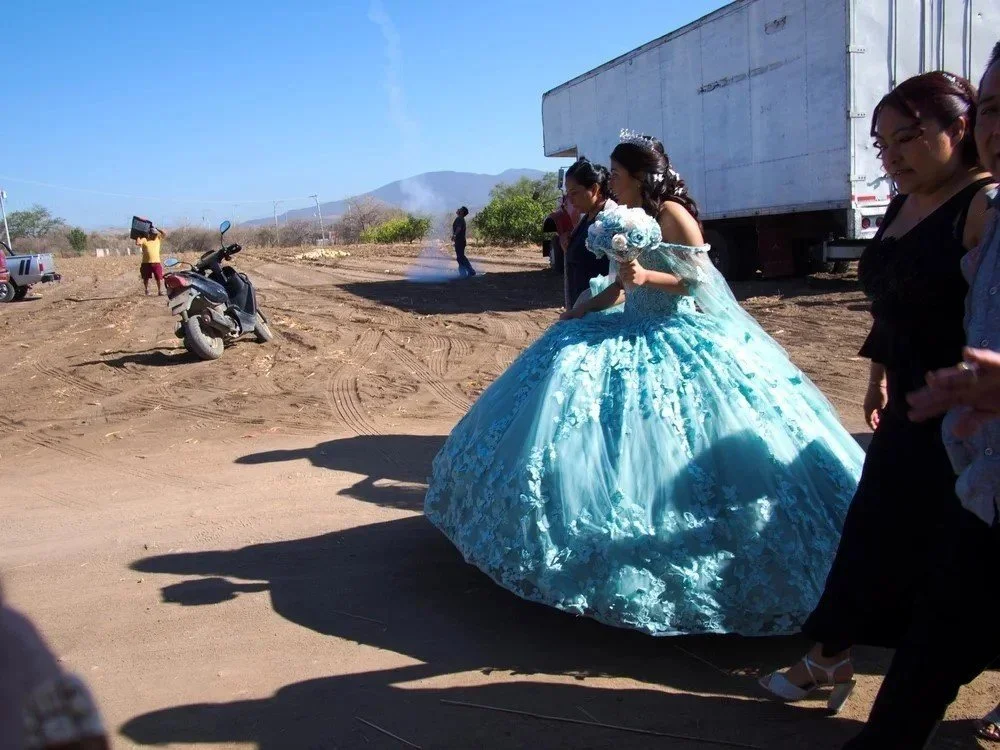 A woman in a blue wedding gown with floral details, holding a bouquet, is walking outdoors beside a group of women. There are trucks, a motorcycle, and mountains in the background on a dirt road.