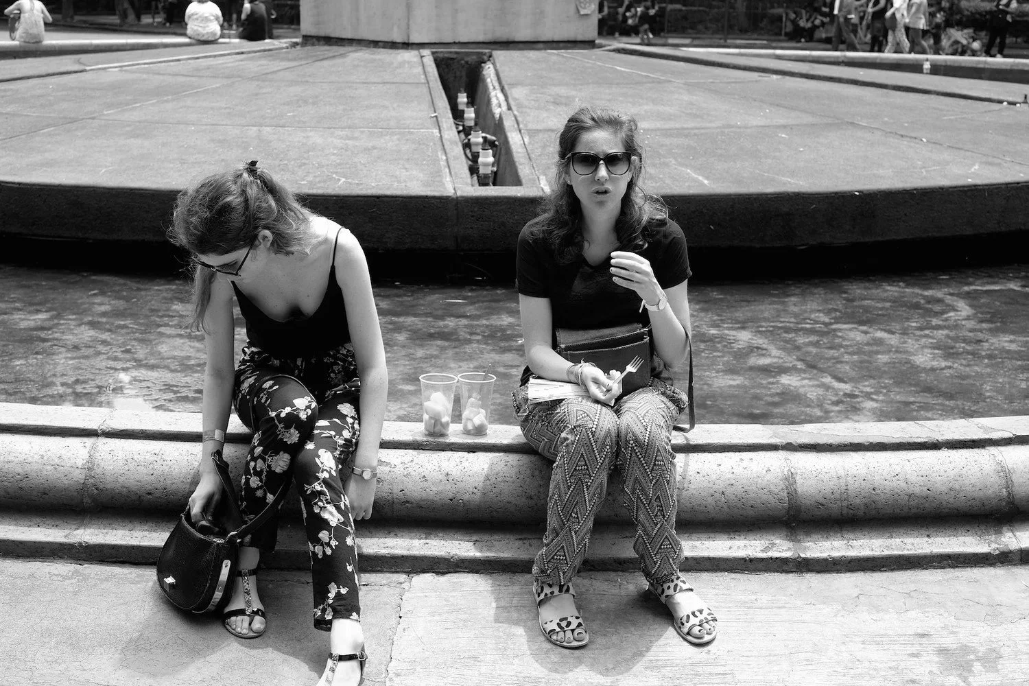 Two women sitting on the edge of a fountain in an outdoor public space, with one looking down and the other looking at the camera, surrounded by a busy area with people in the background.