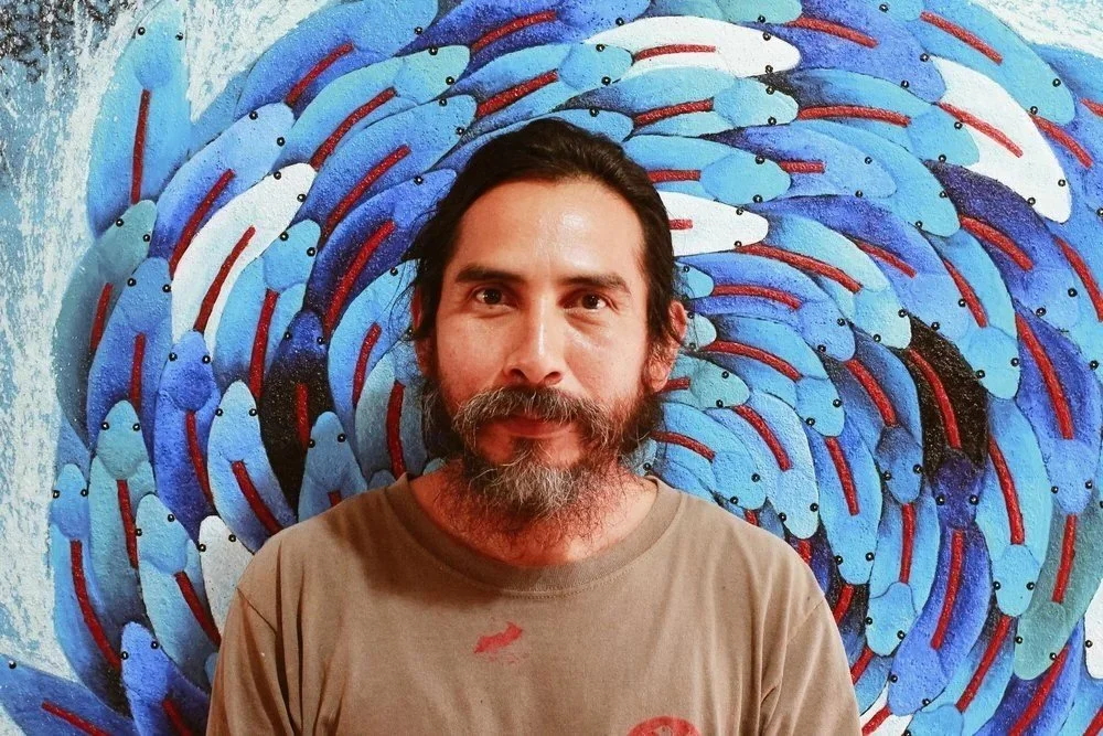 Man with dark hair and beard standing in front of a colorful abstract fish artwork with numerous blue fish swirling around.