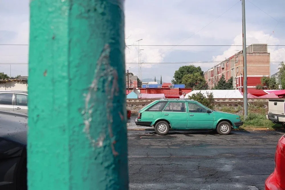 A teal station wagon parked on a cracked asphalt lot with buildings and power lines in the background.