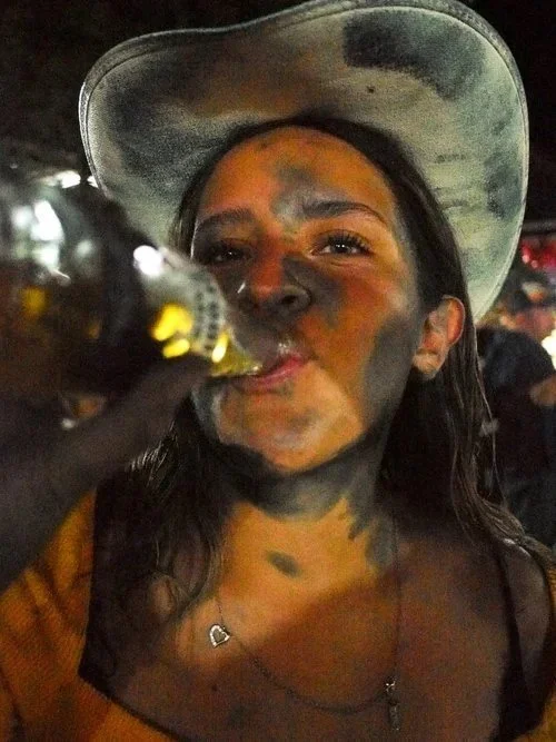 A woman with a sunhat drinking from a bottle, covered in dirt or mud at a nighttime event.