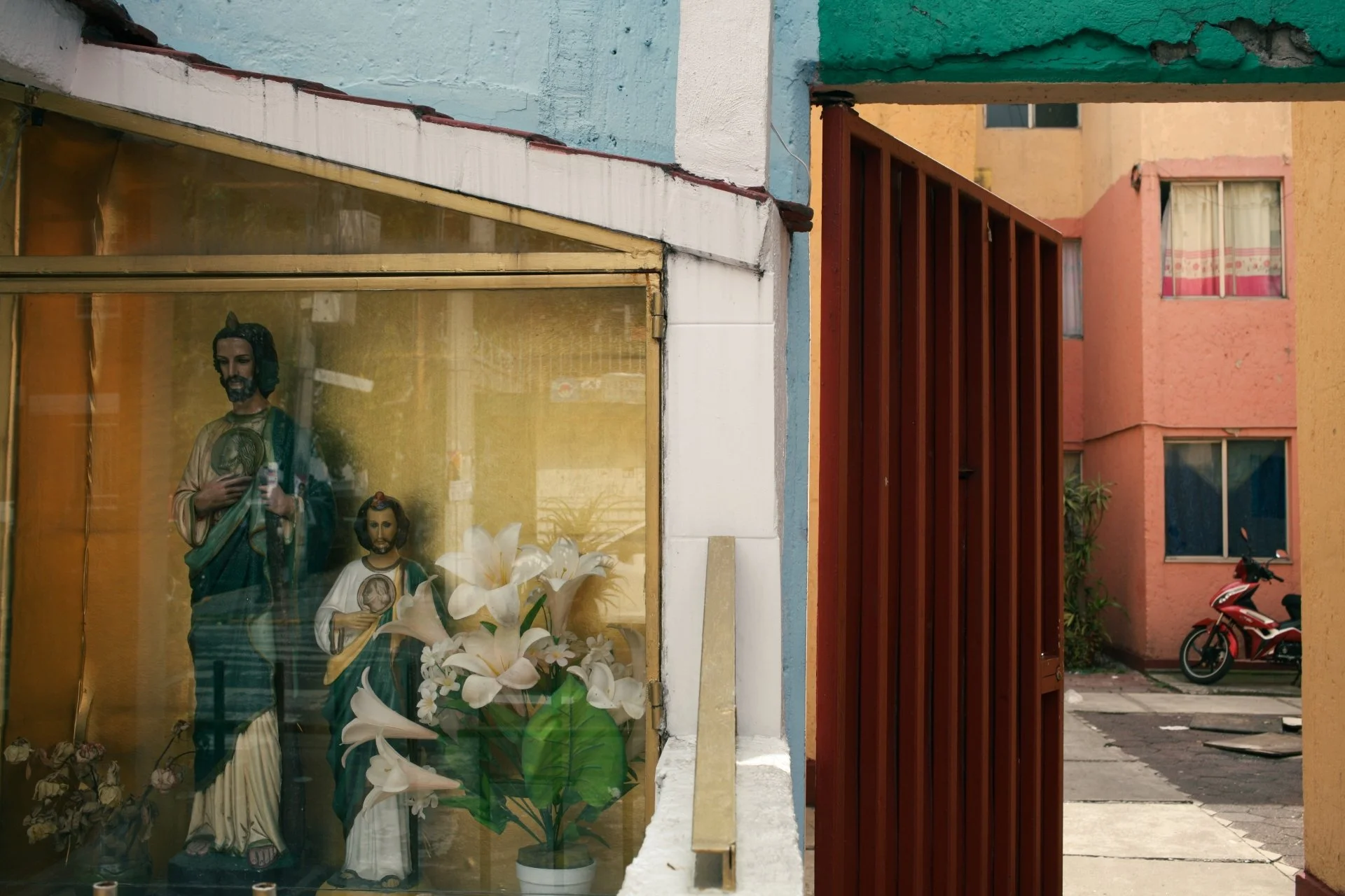 Storefront window display with religious statues of Jesus Christ, a flower bouquet, and some plants inside, with residential building and parked motorcycle outside.