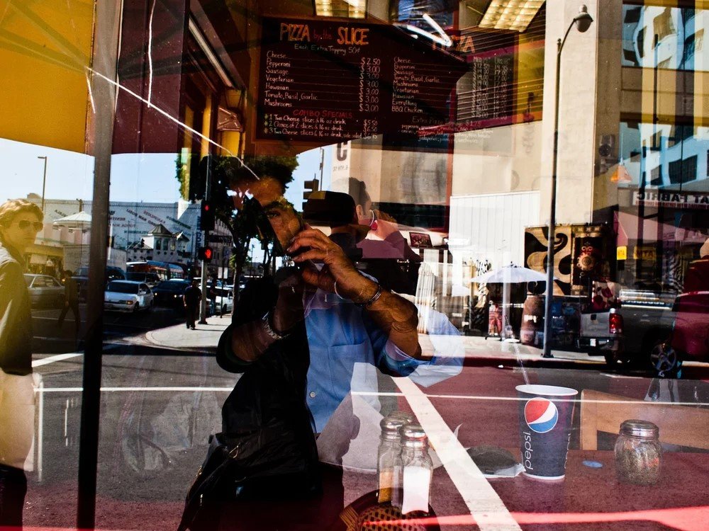Reflection of a person taking a photo inside a restaurant window, with a street scene, including cars and pedestrians, visible outside.
