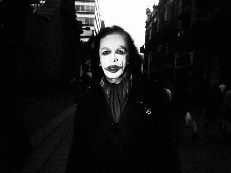 A person dressed as a mime or performer with face paint resembling a clown, standing on a city street at dusk or nighttime.