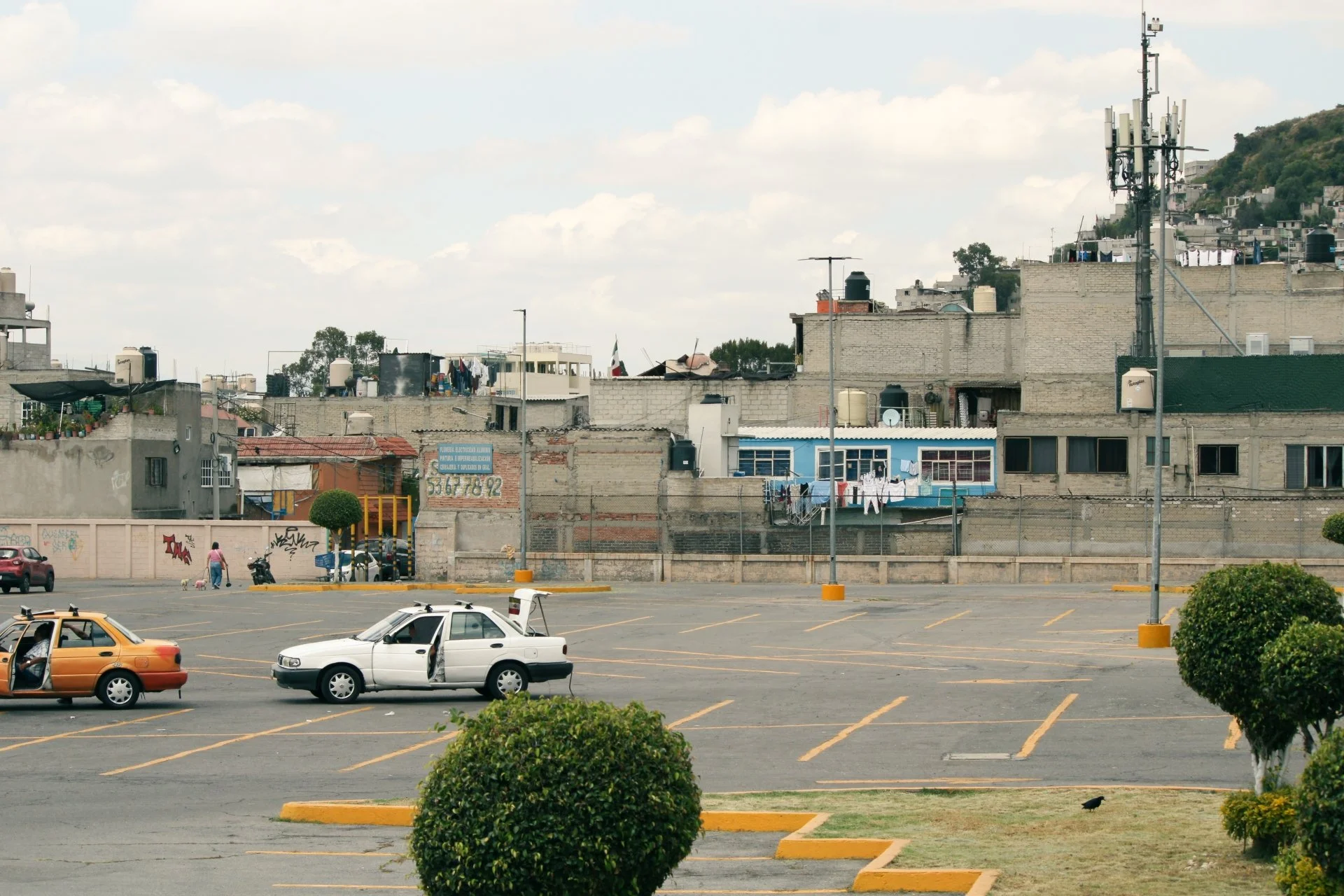 A parking lot in front of a hillside neighborhood with brick and concrete houses, some with laundry hanging outside. Several cars are parked, and a few are driving. There are power lines and antennas on the rooftops.
