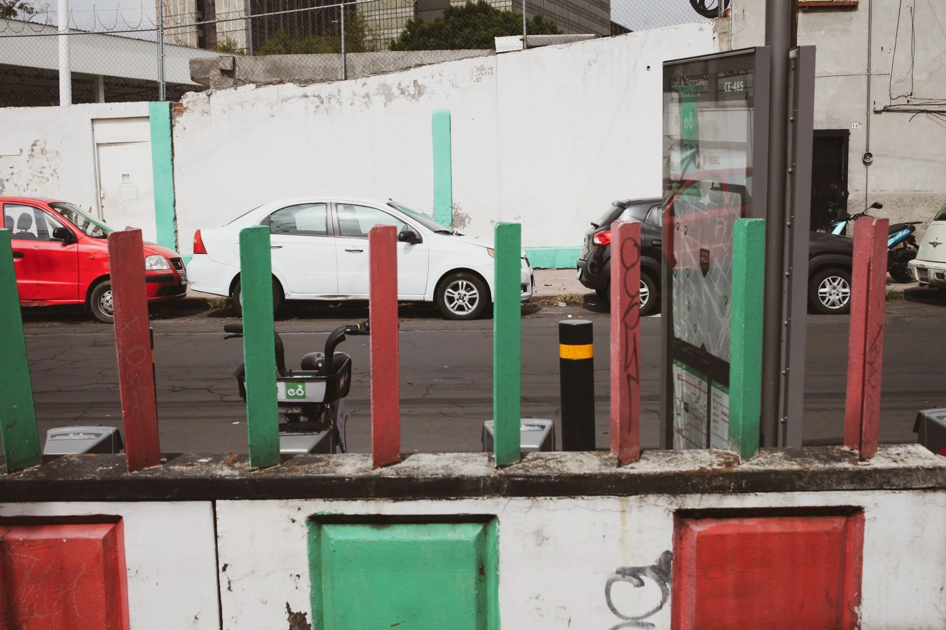 View from a sidewalk through colorful metal bars and a glass bus stop with a map, showing parked cars including a white sedan, a red car, and a black car in front of a white wall with peeling paint and a green painted section. There is a bike-sharing