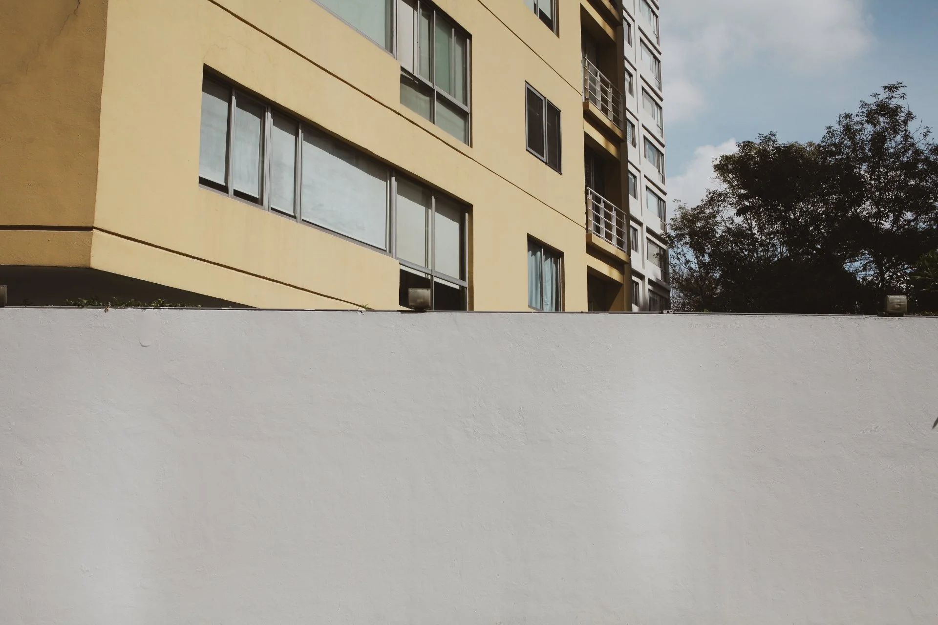 A multi-story yellow apartment building with large glass windows and small balconies, partially obscured by a white wall in the foreground. There are trees on the right side and a partly cloudy sky above.