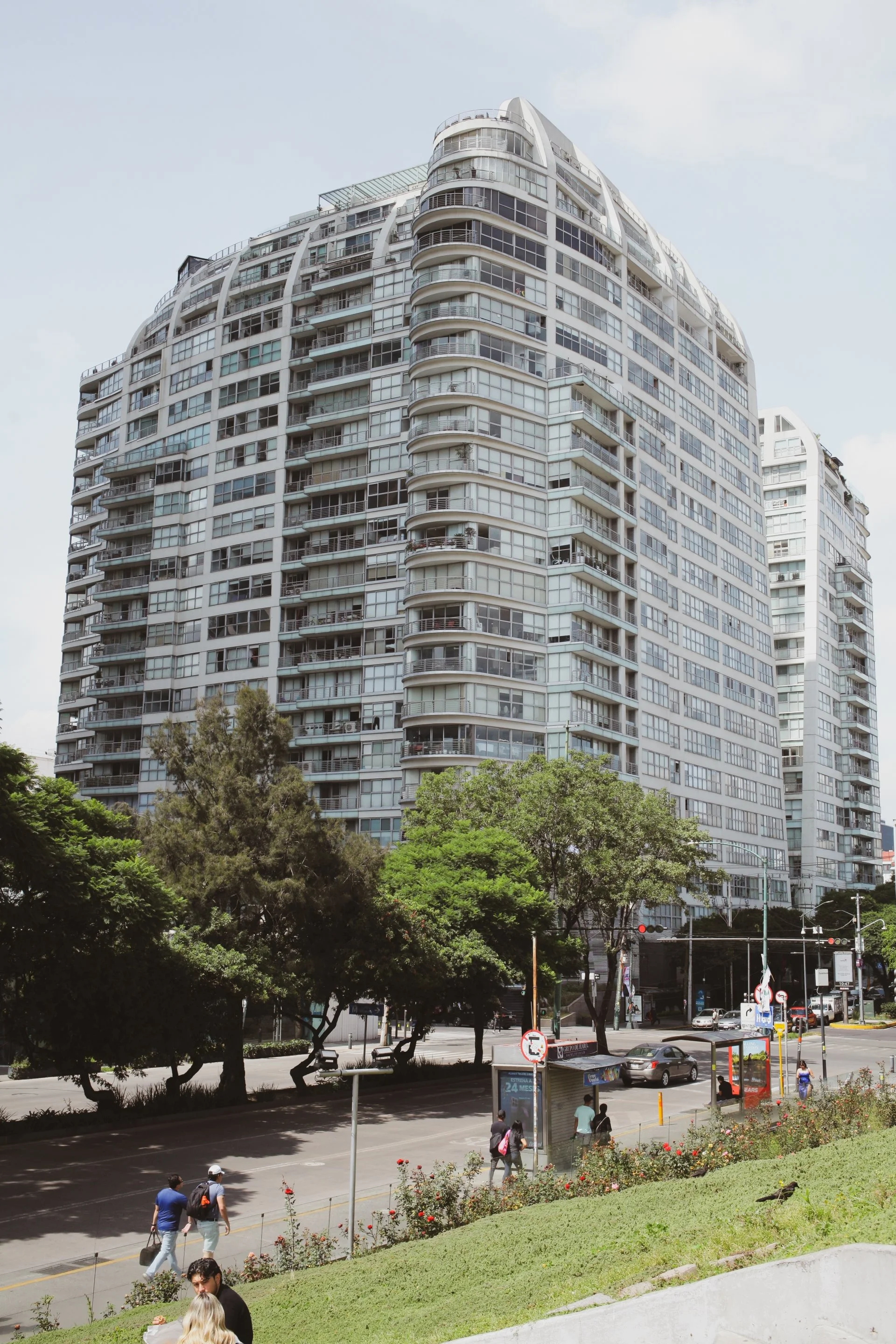 Tall modern residential building with multiple balconies, glass windows, and curved architectural design, seen from street level with trees, people, and vehicles in the foreground.