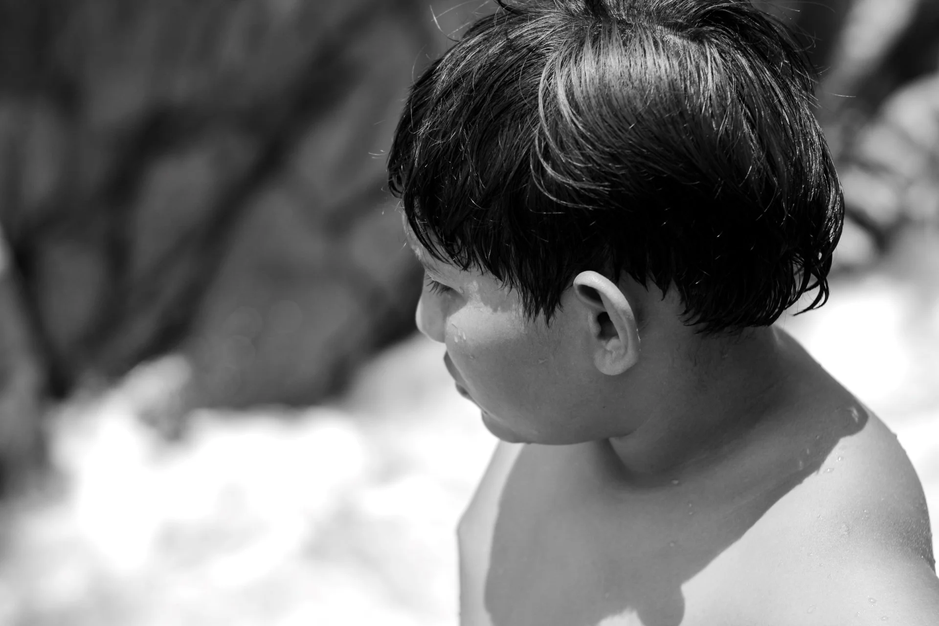 A black and white photo of a young boy with wet, dark hair looking downward, with water droplets visible on his face and neck, outdoors with blurred background.