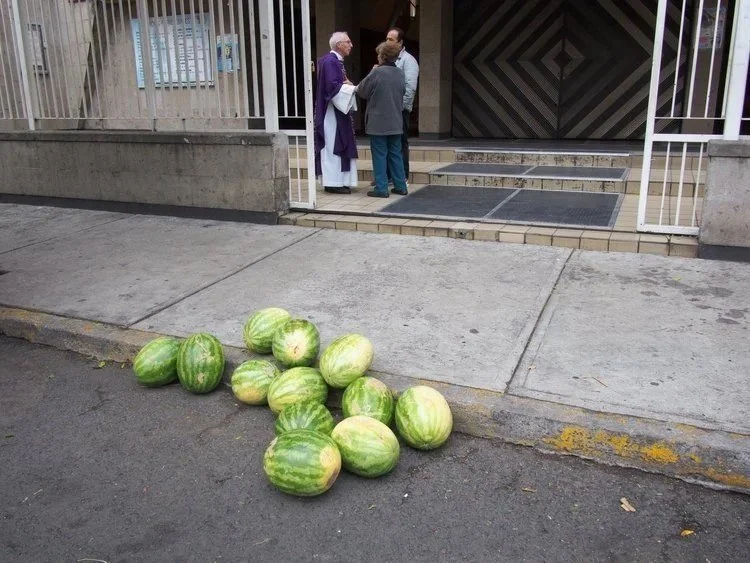 Watermelons are placed on the sidewalk near the curb in front of a building as three people stand and converse at the entrance.