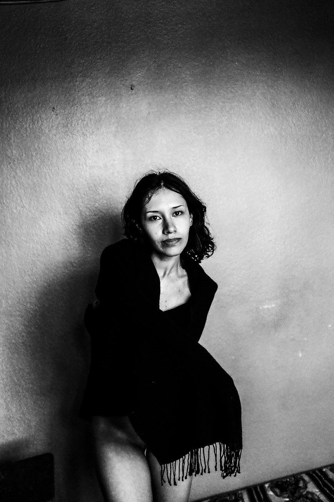 Black and white photo of a woman with curly hair sitting against a plain wall, wearing a dark dress with fringes at the bottom.