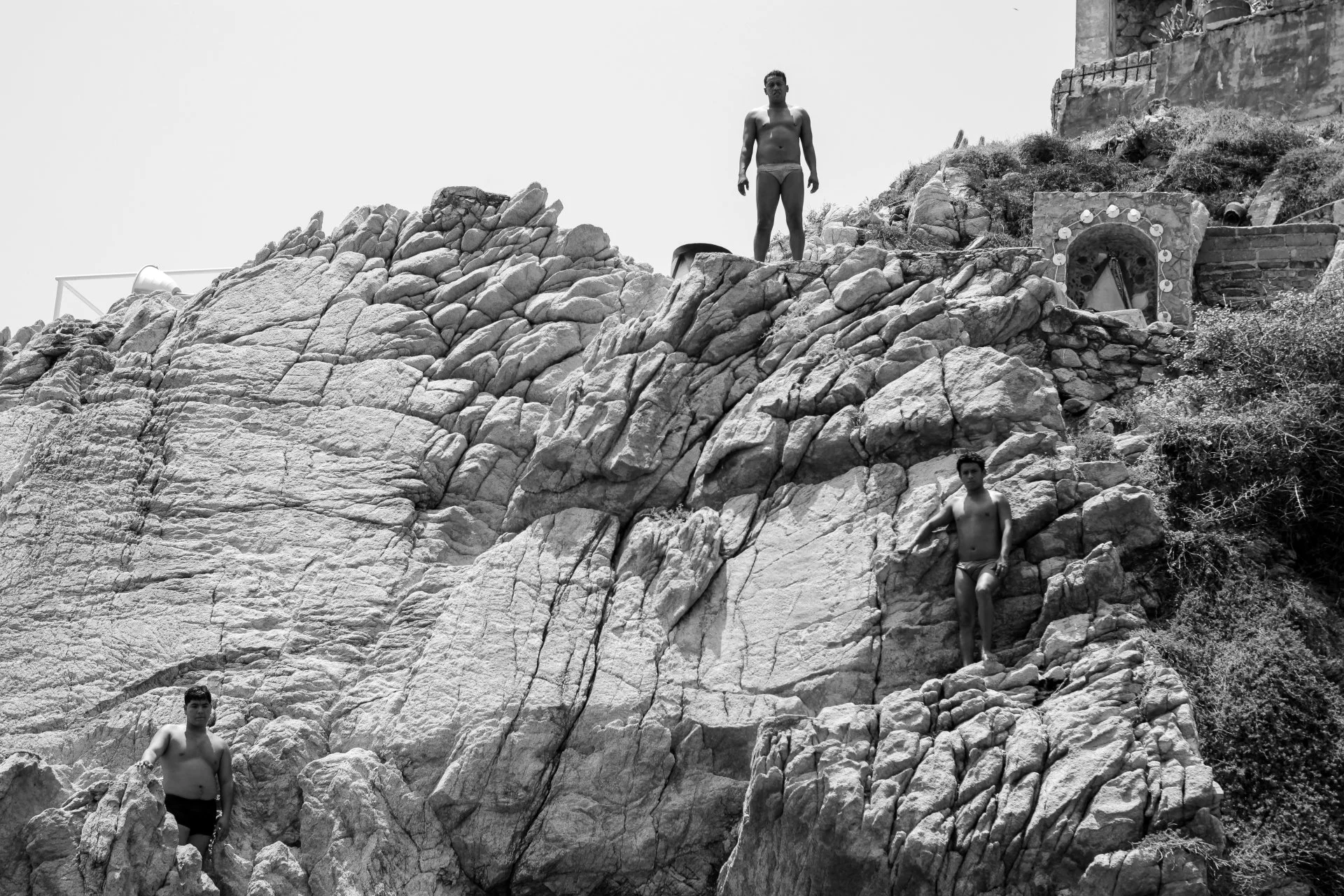 Three young men, shirtless and wearing swim trunks, are on rocky terrain near the coast. One is standing on higher rocks, another is sitting lower, and the third is partially obscured at the bottom left.