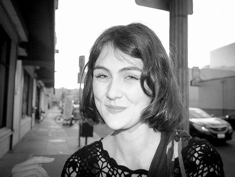 A woman with short dark hair and a patterned top winks and smiles at the camera on a city street.