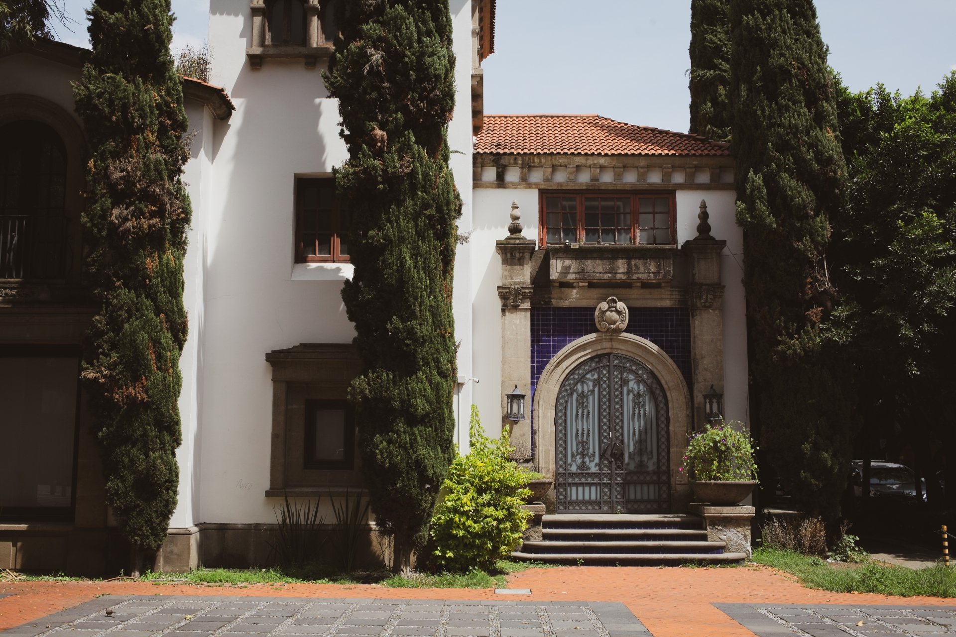 The image shows the entrance of a historic building with an ornate stone archway and decorative urns on top. The building has a red clay tile roof and tall, narrow windows. Two tall cypress trees flank the entrance, and a stone pathway with stairs le
