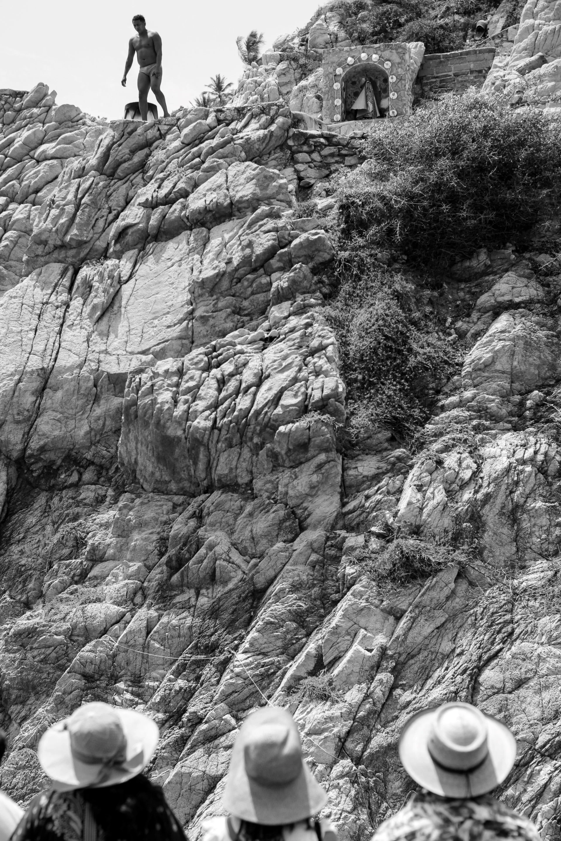 A group of people wearing hats seen from behind, observing a woman walking along a rocky slope at a mountain or hill.