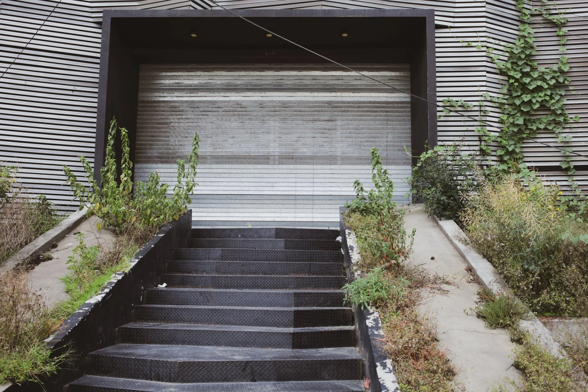 Exterior of a modern building with a black metal staircase leading up to a metal roll-up door, surrounded by plants and greenery, with a striped metal facade.