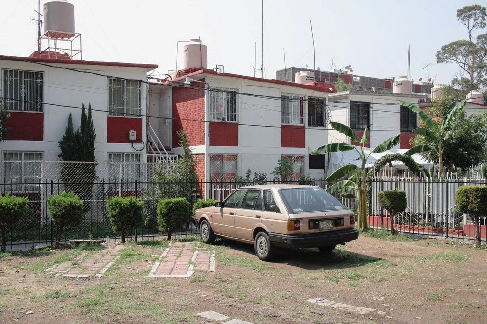 A beige vintage hatchback car parked in front of a fenced residential building with red and white walls, high windows, and tropical plants including banana trees.