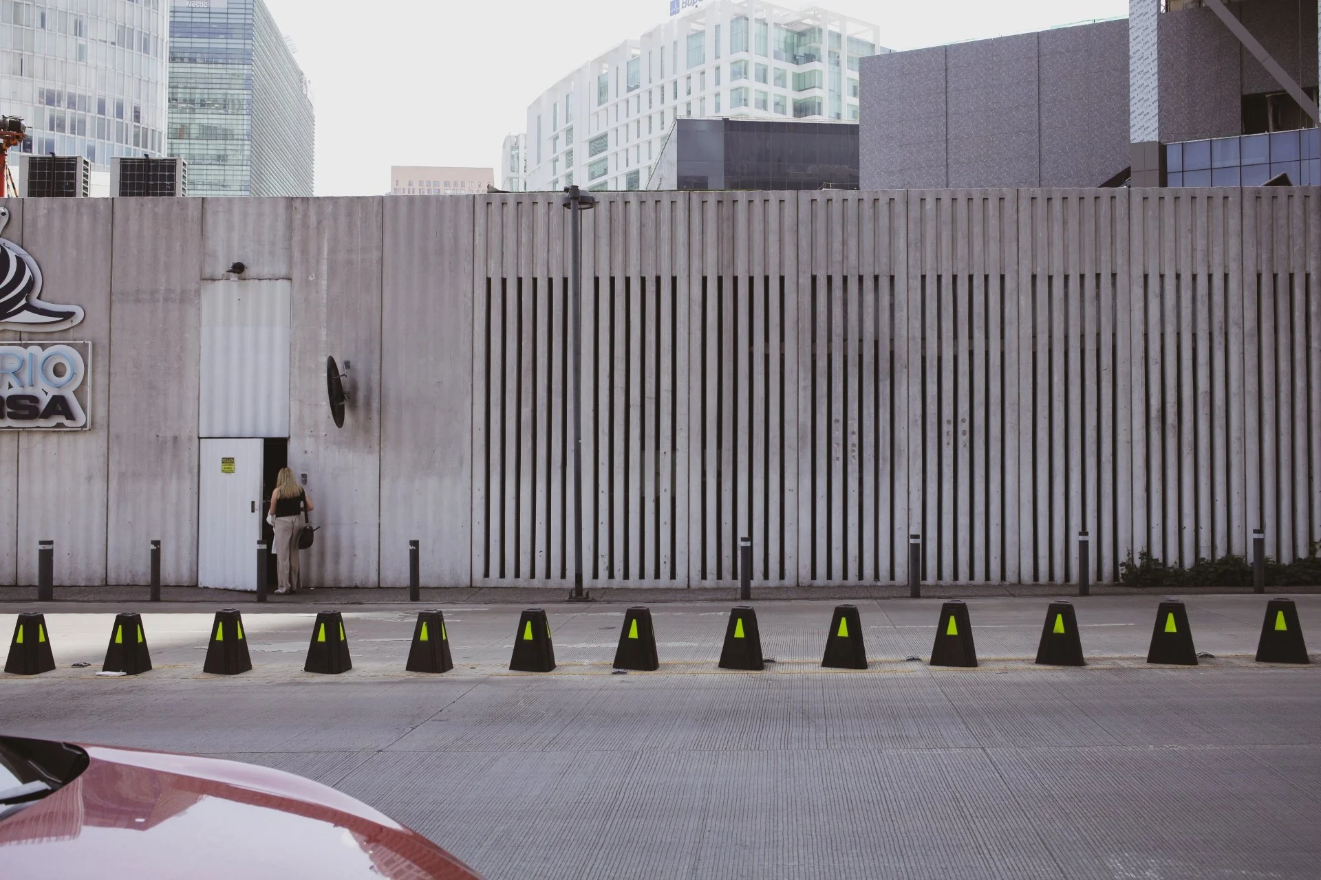 A woman standing on the sidewalk near a concrete wall in an urban area with tall buildings in the background, and black traffic cones in the foreground.