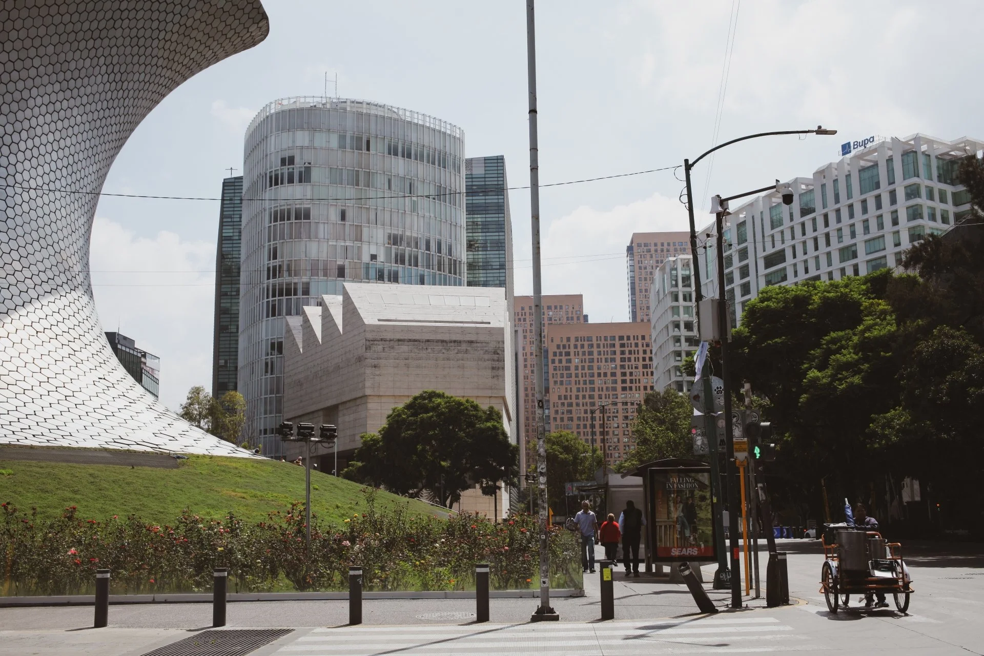 Cityscape of modern skyscrapers with a park and a bus shelter in the foreground, some people walking and a wheelchair near the sidewalk.