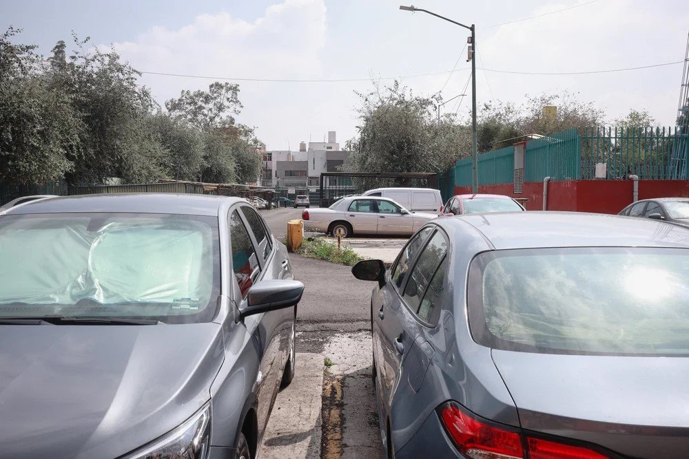 Parking lot with several parked cars, some covered with plastic or fabric, surrounded by trees, buildings, and a fence, under a partly cloudy sky.