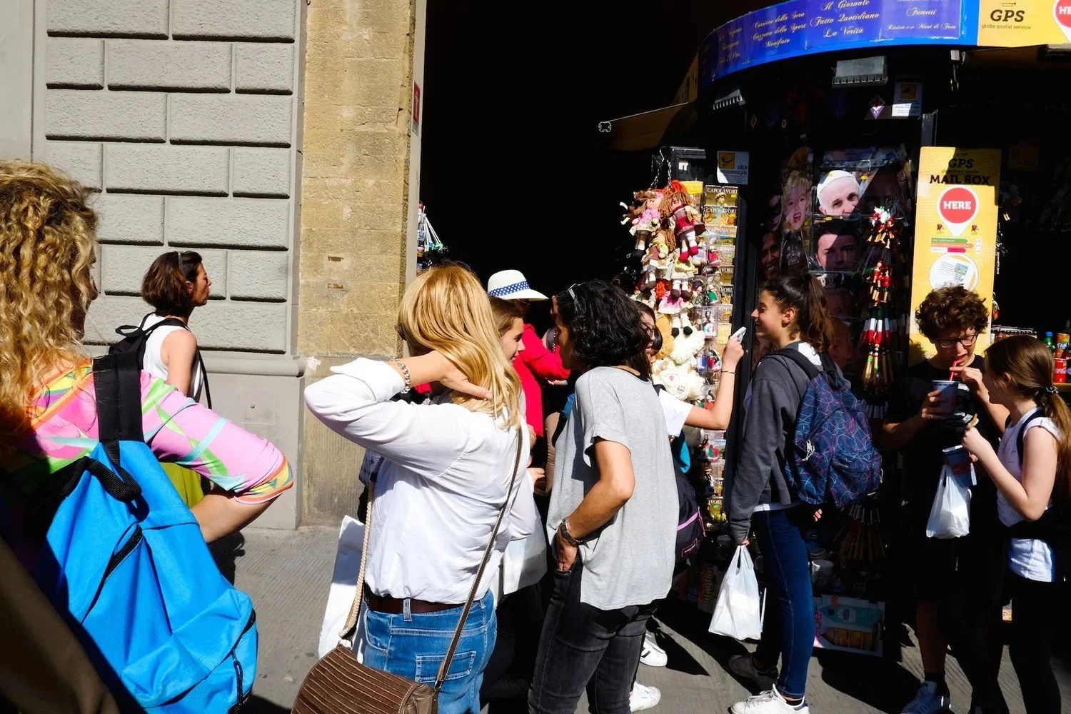 Group of people standing in line on a city sidewalk, chatting and looking at a souvenir shop window. The shop displays plush toys, hats, and postcards. The group is of diverse ages and styles, with some carrying backpacks and shopping bags, dressed i