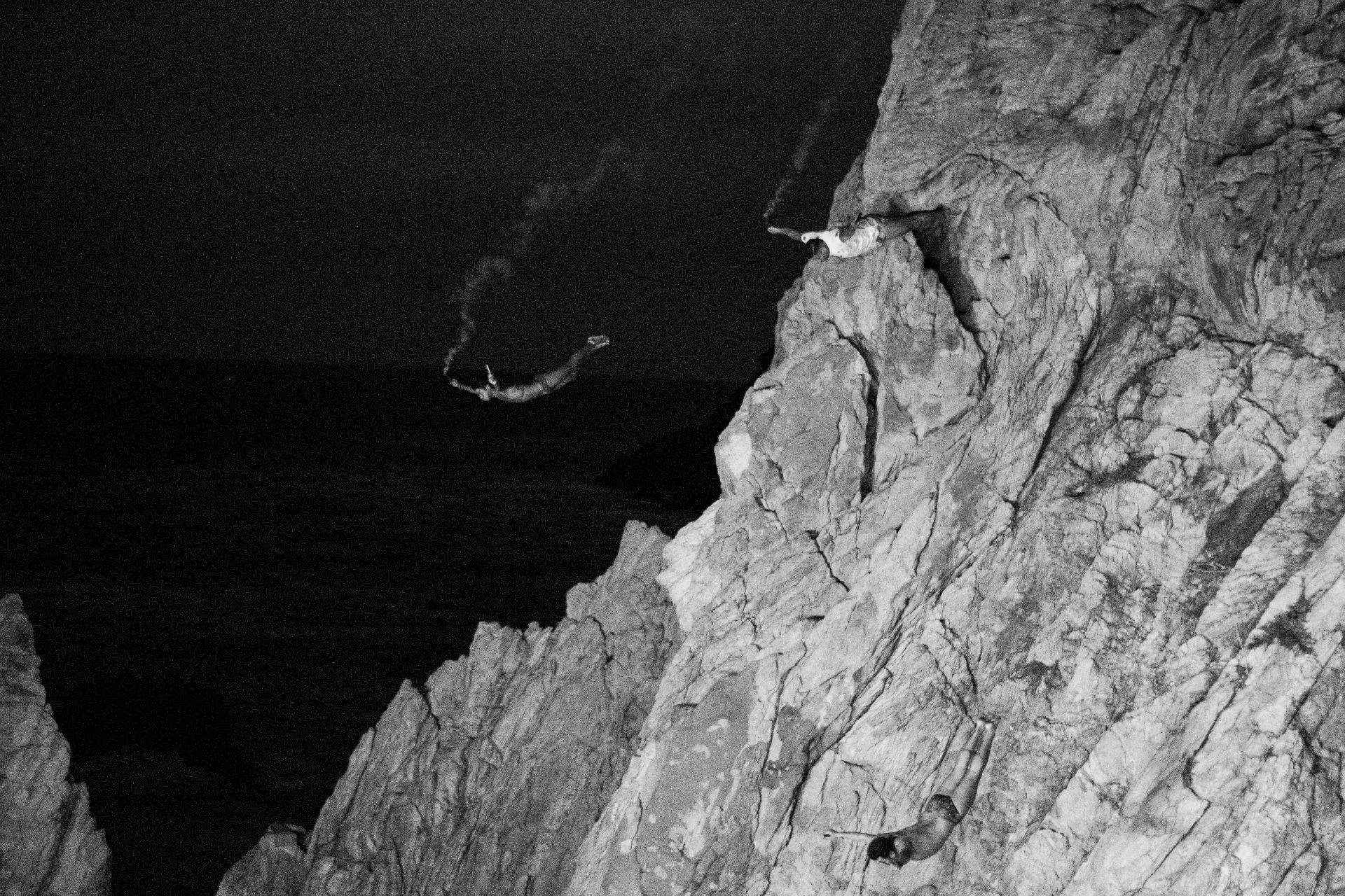 Two individuals are climbing and sitting on rocky cliffs at night, one near the edge looking down, both wearing headlamps.