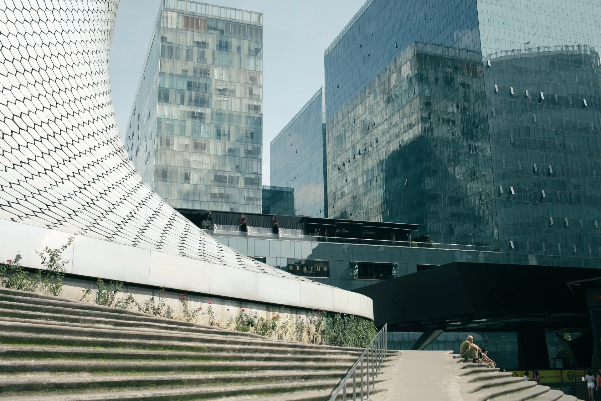 Modern cityscape featuring tall glass and steel buildings, a concrete staircase with a few people sitting on the steps, and a curved architectural structure on the left side of the image.