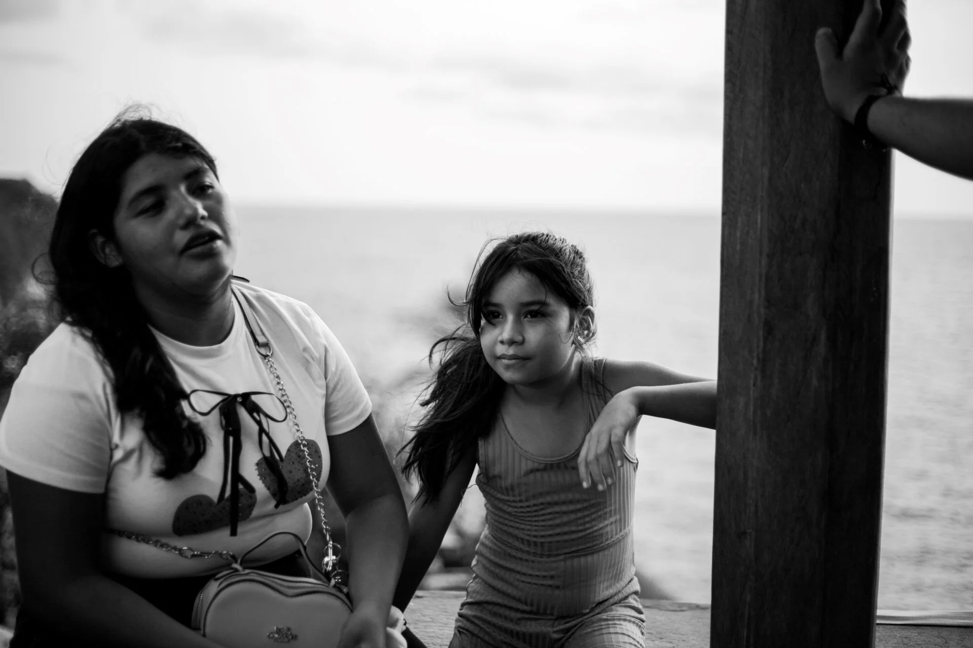 Two young girls, one with long dark hair and the other with lighter hair, sitting outdoors by the water, next to a wooden post, in a black and white photo.