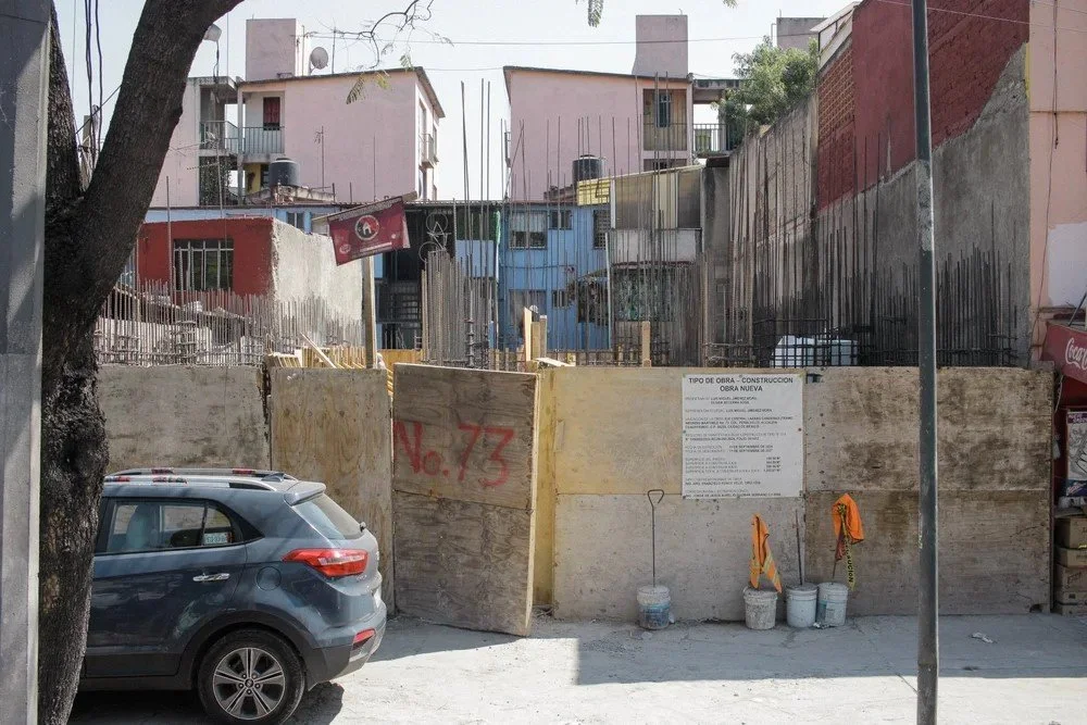 Construction site with a concrete wall, wooden boards, construction signs, buckets, orange safety vests, parked car, trees, and residential buildings in the background.