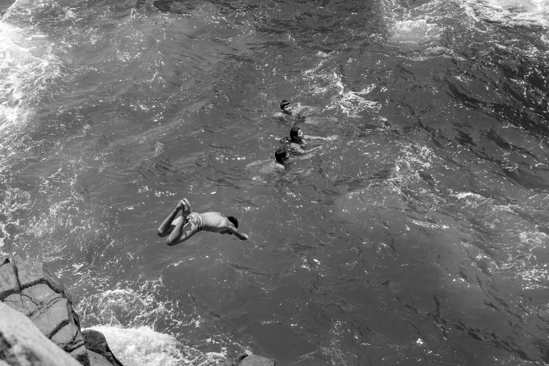 Black and white photo of four children swimming in a body of water, with a rocky edge visible at the bottom left.