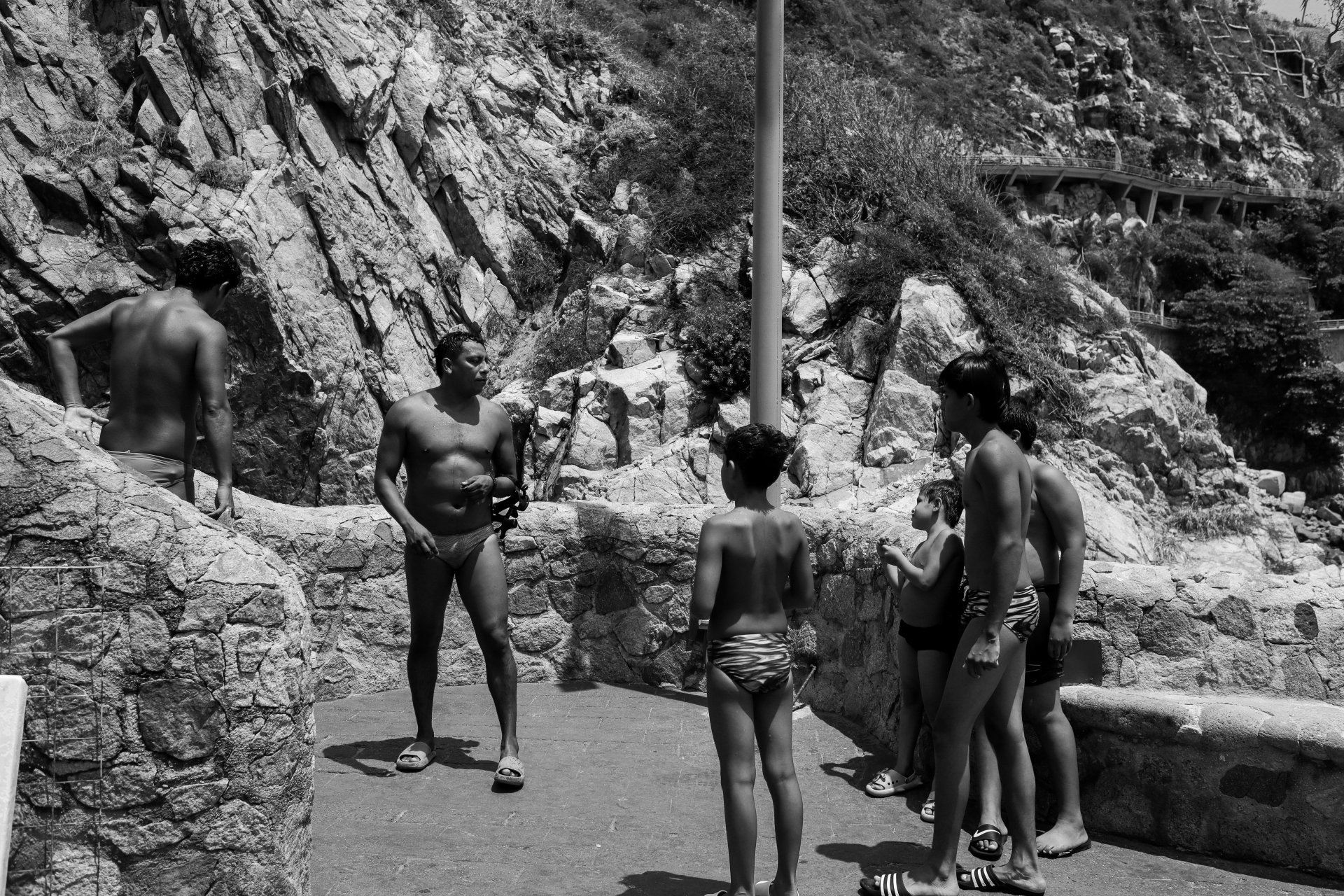 A group of people, mostly children, in swimsuits gathered on a rocky outdoor area with a stone wall and hillside in the background. Some appear to be listening to a man in swim trunks speaking to them.