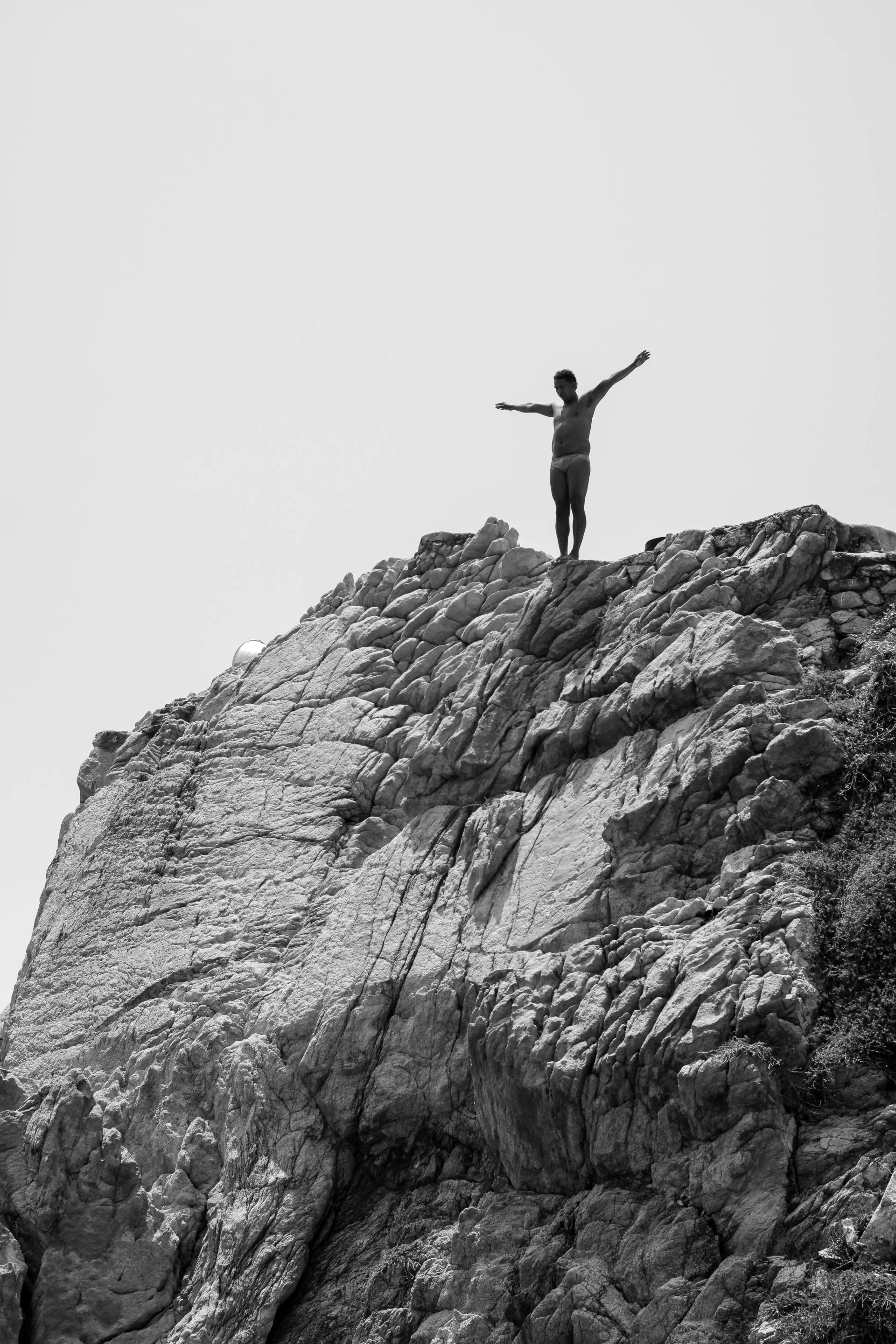 A person standing with arms outstretched on top of a rocky mountain or cliff, high above the ground, in black and white.