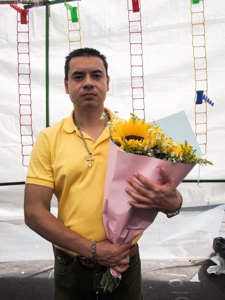 A man wearing a yellow polo shirt holding a bouquet of yellow flowers wrapped in pink paper, standing in front of a white background with colorful plastic ladders hanging.