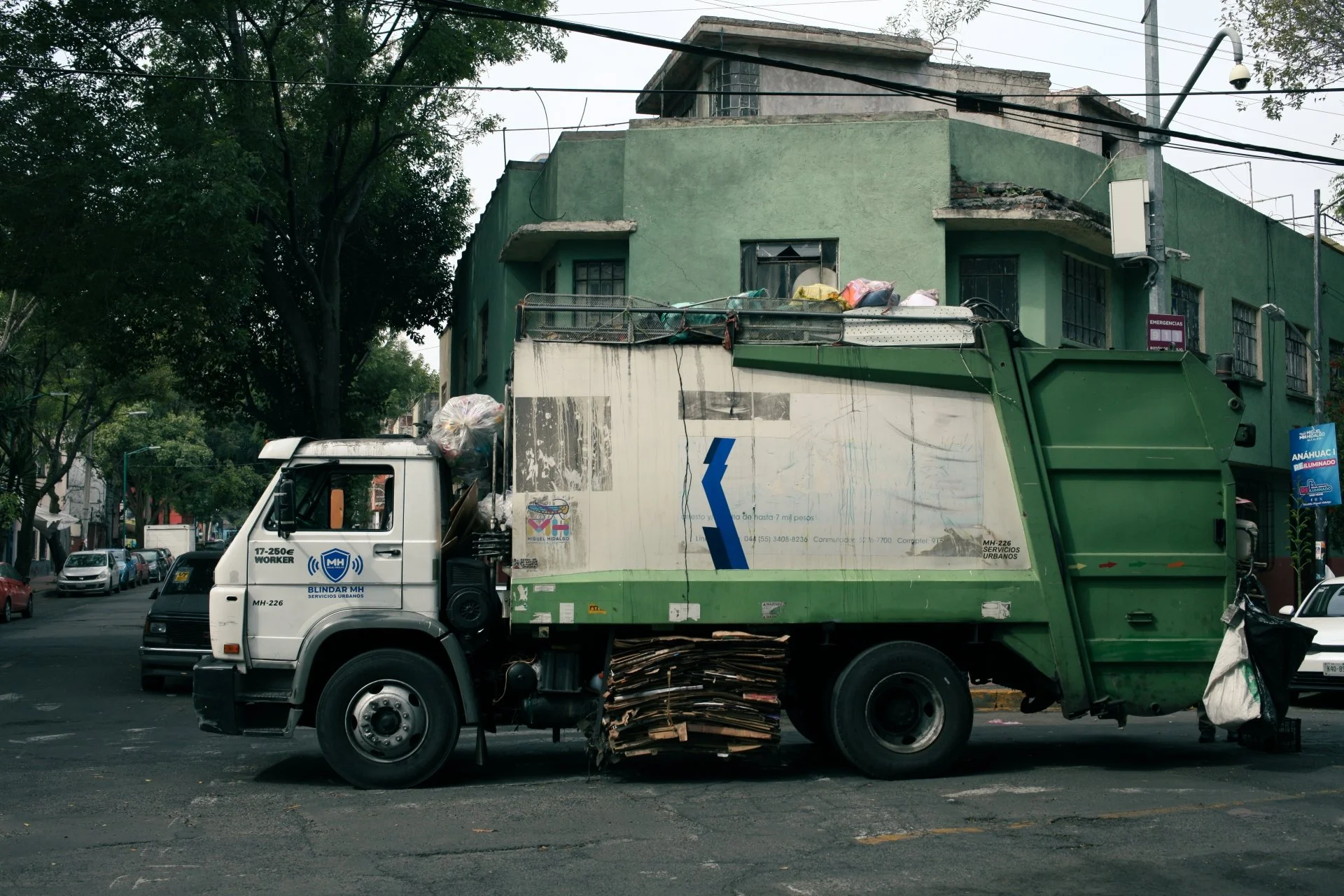 A green and white trash collection truck parked on a city street with a green building in the background. The truck is loaded with trash and cardboard.