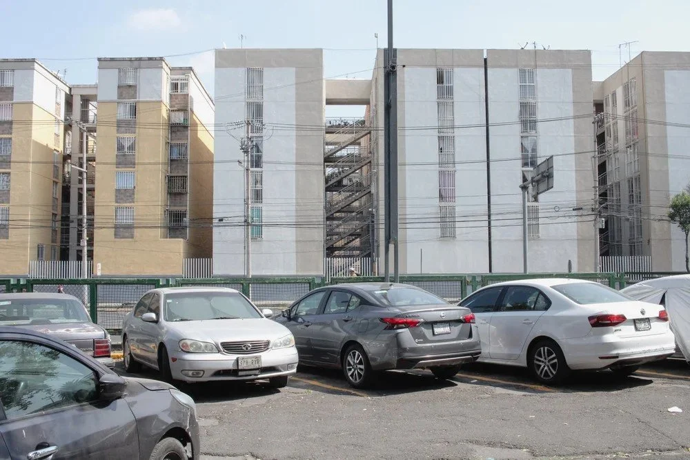 A parking lot filled with cars in front of a large apartment building with beige and white walls, surrounded by electrical wires.