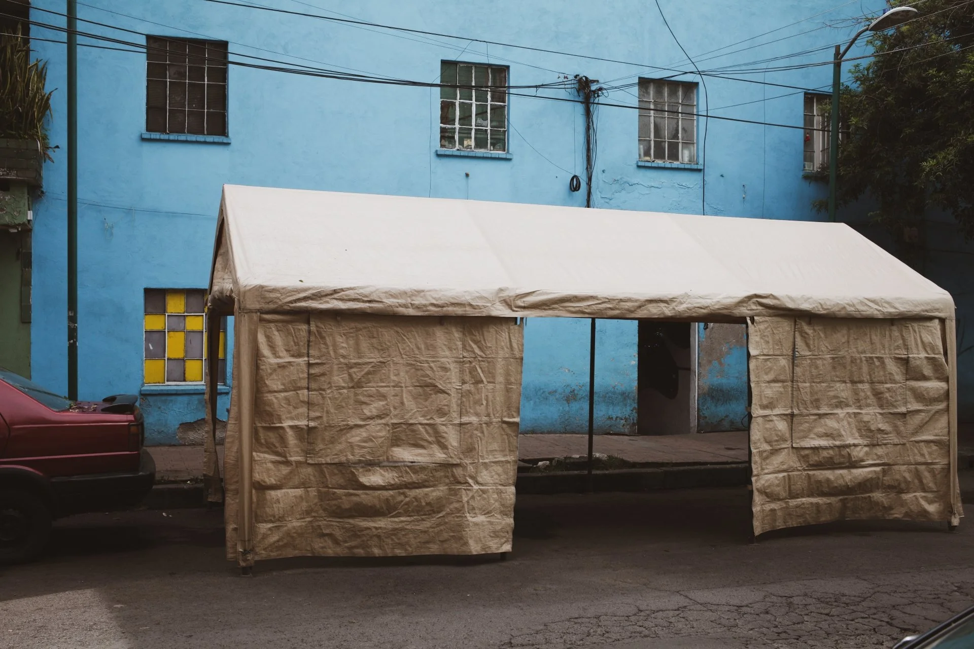 A beige canopy tent set up on a city street with a blue building in the background, along with intersecting electrical wires.
